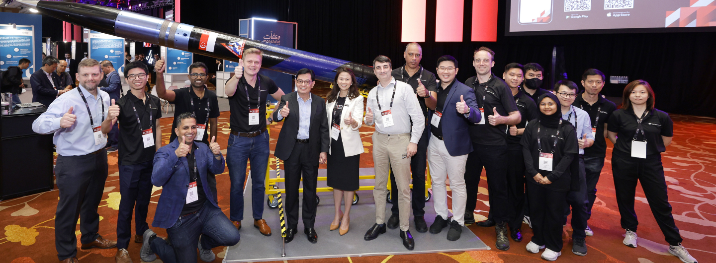 Group of people giving thumbs up in front of a large rocket with a Singaporean flag.